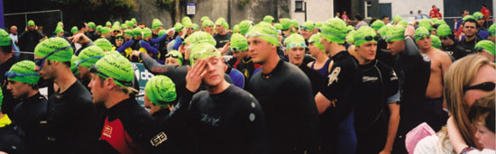 a crowd of TriAthletes mill about in their green swim hats, goggles and wet suits on the banks of the river shannon in athlone